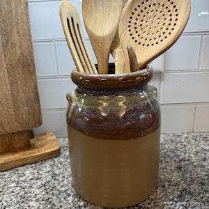 Puede incluir: Un recipiente de cerámica marrón y beige para utensilios, lleno de cucharas y espátulas de madera. El recipiente tiene un borde esmaltado y una forma cilíndrica, ideal para la cocina.