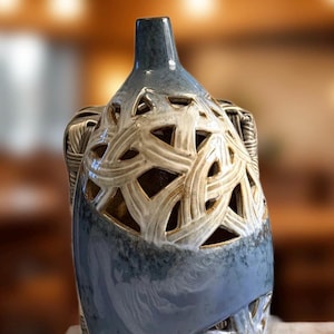May include: A decorative ceramic vase with a blue and cream color scheme. The vase features a woven pattern with cut-out sections. The top and base are a cream color, while the middle section is blue. The vase is approximately 12 inches tall.