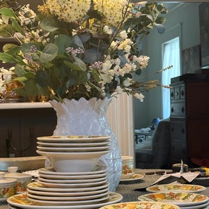May include: A collection of cream-colored ceramic dinnerware featuring a colorful fruit pattern along the rims. The set includes stacked dinner plates, salad plates, and bowls, arranged in front of a white textured vase filled with faux flowers.