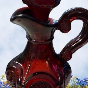 May include: A ruby red glass pitcher with a handle and a stopper. The pitcher has a decorative pattern on the body and handle. The stopper is a rounded shape with a textured surface. The pitcher is likely for serving beverages.