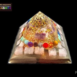 May include: A pyramid-shaped orgonite with a gold-colored top and base, filled with crystals and colorful beads. The pyramid has a clear resin exterior and the text "OM ORGONITE" in the upper left corner.