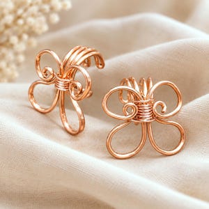 May include: Two rose gold-colored wire ear cuffs shaped like stylized butterflies. Each piece is crafted from a single continuous length of metal wire, featuring delicate loops and a central coiled section that secures the cuff to the ear cartilage.