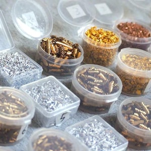 Könnte beinhalten: Eine Sammlung kleiner, klarer Plastikbehälter, gefüllt mit verschiedenen metallischen Komponenten, darunter Schrauben und Granulate in Gold-, Silber- und Kupfertönen. Die Behälter sind auf einer hellen Oberfläche angeordnet. Der Text "OM Orgonite" ist sichtbar.