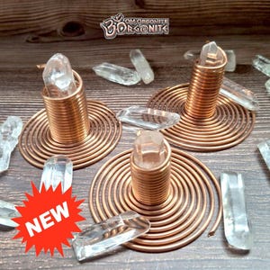 Könnte beinhalten: Orgonit-Stücke aus Kupferdraht mit Bergkristallen. Jedes Stück hat eine spiralförmige Basis und eine zentrale Spule, die einen Kristallpunkt hält. Das Wort "NEW" wird auf einem roten Stern angezeigt. Das OM Orgonite-Logo ist im Hintergrund.