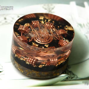 May include: A round, clear resin orgonite with copper coils and gold flakes. The orgonite has a dark base and a clear top, with the text "OM ORSONITE" at the top left and "TB 112" at the bottom right.