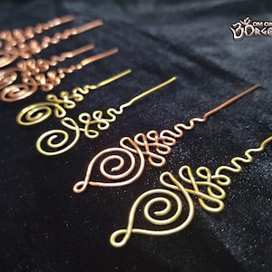 Könnte beinhalten: Eine Sammlung handgefertigter Haaraccessoires aus Draht in verschiedenen Metallausführungen, darunter Kupfer und Gold. Die Designs weisen komplizierte Wirbel und dekorative Elemente auf. Das Bild enthält den Text "OM ORGONITE".