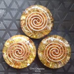 Può includere: Tre pezzi rotondi di orgonite con un design a spirale in rame. I pezzi sono riempiti di scaglie dorate e marroni. Il testo "OM Orgonite PD159" è in basso.