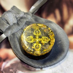 May include: A round, clear orgonite piece with a gold-colored design, set in a gray velvet pouch. The orgonite has gold flakes and a gold-colored symbol. The text "Om Orgonite" is visible on the pouch.