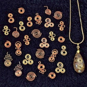 May include: Various copper-coloured wire spirals and decorative elements are scattered on a dark surface. A gold-toned necklace with a teardrop pendant, also featuring a spiral design, is visible. The jewellery components vary in size and shape.