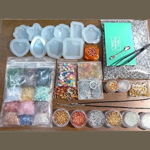 May include: A craft kit with various supplies for jewelry making. Includes silicone molds in different shapes, bags of colorful beads and stones, containers of glitter, and metal findings. A green card with text and a silver spoon are also included.