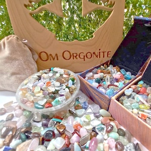 Puede incluir: Una variedad de gemas y cristales de colores se exhiben en un cuenco transparente, cajas abiertas y dispersos sobre una superficie. Un recorte de madera con las palabras "Om Orgonite" está en el fondo, junto con una pequeña bolsa beige.