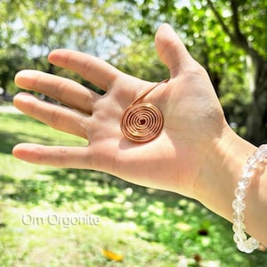 May include: A copper wire spiral ornament featuring a circular coil design. The metallic copper piece is shaped into a flat, concentric spiral with a single wire tail extending from the outer edge. The text Om Orgonite is visible in the corner.