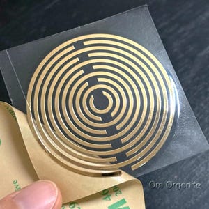 Può includere: Un adesivo circolare color oro caratterizzato da un motivo a spirale concentrica con un'apertura centrale. L'adesivo è parzialmente staccato dalla carta di supporto color beige, rivelando uno strato adesivo trasparente. Il testo Om Orgonite è visibile.