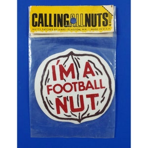 May include: A vintage patch in a clear plastic package. The patch is white with a brown outline and features the text "I'M A FOOTBALL NUT" in red. Above the patch, a yellow banner reads "CALLING ALL NUTS!"