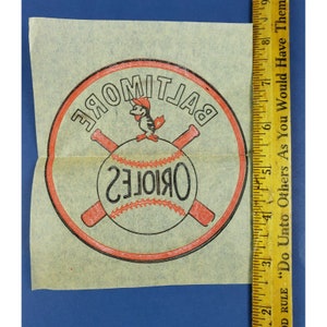 May include: A vintage Baltimore Orioles baseball logo sticker with a red outline and a black and white illustration of a bird holding a baseball bat. The text "Baltimore Orioles" is written in a circular pattern around the bird.