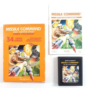 May include: A vintage Atari Missile Command video game set including an orange cardboard box, an instruction manual, and a black game cartridge. Each item features the game title and artwork depicting a person operating a missile defense system.
