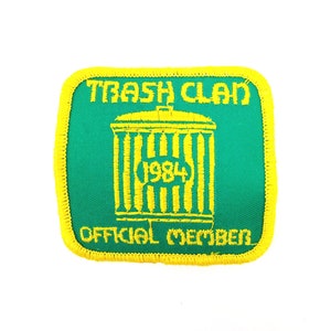May include: A green and yellow embroidered patch with the text "Trash Clan 1984 Official Member". The patch features a yellow trash can with a yellow lid.
