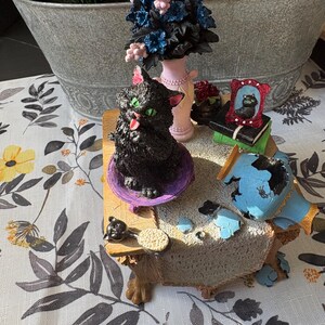 May include: A whimsical figurine set featuring a black cat with green eyes, a pink-colored vase with blue and pink flowers, and a miniature framed picture of a cat. The set is on a decorative table with a floral tablecloth.