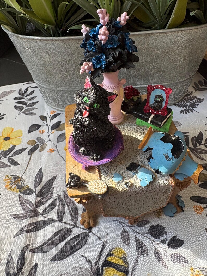 May include: A whimsical figurine set featuring a black cat with green eyes, a pink-colored vase with blue and pink flowers, and a miniature framed picture of a cat. The set is on a decorative table with a floral tablecloth.