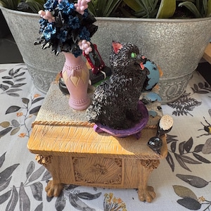 May include: A decorative figurine featuring a black cat holding a globe, seated on a small wooden table. A pink vase with blue and pink flowers sits beside the cat. A black telephone and other small items are also on the table.