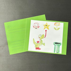 Könnte beinhalten: Eine grüne Grußkarte mit einer Aquarellillustration eines Zeichentrickdinosauriers, der nach einem Stern greift. Die Karte zeigt auch einen Pilz, eine Blume und ein grünes Rohr. Die Karte ist geöffnet und zeigt das Innere.
