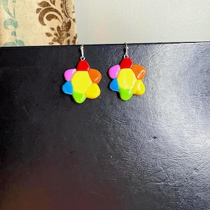 Op de afbeelding: Een paar kleurrijke bloemvormige oorbellen. Elke oorbel heeft bloemblaadjes in rood, oranje, geel, groen, roze en blauw. De oorbellen hangen aan zilverkleurige haken, wat een speels en opvallend accessoire biedt.