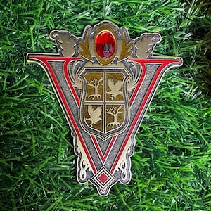 May include: A decorative pin in the shape of a V, featuring a crest design with a red gem at the top. The crest includes trees and birds, with a red and gold border. The pin is set against a backdrop of green grass and red rose petals.