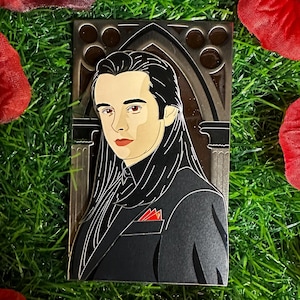 May include: A rectangular enamel pin featuring a stylized portrait of a person with long black hair and a black suit with a red pocket square. The pin is set against a backdrop of green grass and surrounded by red rose petals.