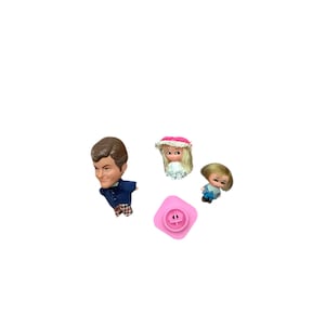 May include: A collection of vintage doll heads and a pink plastic toy. The doll heads include a man with brown hair and a blue jacket, a girl with a pink hat, and a blonde-haired doll. The pink toy is square with a circular design.
