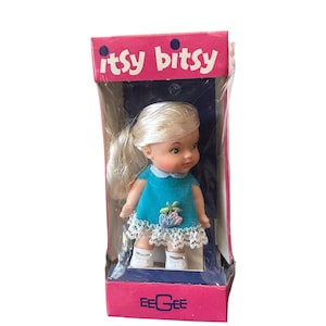 May include: A small doll with blonde hair in a teal dress with a white lace hem and floral embroidery, packaged in a pink box labeled itsy bitsy and EEGEE. The doll wears white shoes and has a light skin tone, presented in a clear plastic sleeve.