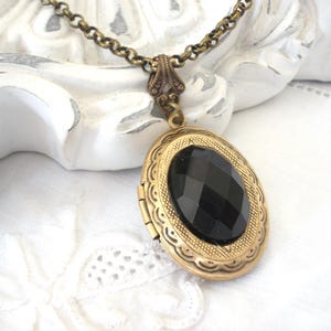 Op de afbeelding: Een gouden ketting met een hanger in de vorm van een medaillon, bezet met een zwarte, gefacetteerde edelsteen. Het medaillon heeft een versierd ontwerp en is aan een ketting bevestigd.
