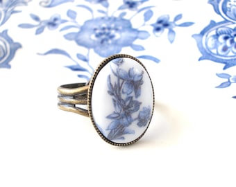 May include: A ring with a white oval ceramic top featuring a blue floral design. The ring band is made of brass with a triple band design.