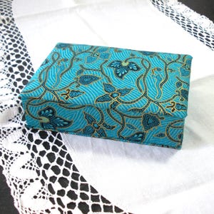 May include: A rectangular box with a turquoise batik pattern featuring floral and vine motifs in black and gold. The box is set on a white lace-trimmed surface, creating a contrast of textures and colours.