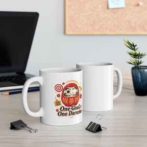 May include: White ceramic mug with a Daruma doll design and the text "One Goal, One Daruma." The Daruma doll is red with gold accents and a target symbol. The mug has a white handle and a glossy finish.