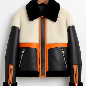 May include: A stylish jacket with a cream-coloured body, black sleeves, and a black collar. The jacket features orange accents and zips. The jacket is hanging on a wooden hanger.