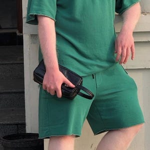 May include: A person wearing a green t-shirt and shorts set, holding a black clutch. The outfit has a raw hem. The clutch is rectangular with a handle.