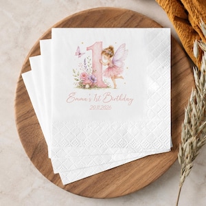 May include: A stack of white paper napkins featuring a watercolour illustration of a pink number one, a fairy with wings, flowers, and a butterfly. The text reads Emma's 1st Birthday and 20.11.2026 in a pink script font with a textured embossed border.