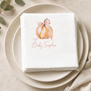 May include: A stack of white paper napkins featuring a watercolour illustration of an orange pumpkin tied with a light pink ribbon. Below the pumpkin, the text Baby Sophia is printed in a cursive pink font. The napkins are square with a textured edge.