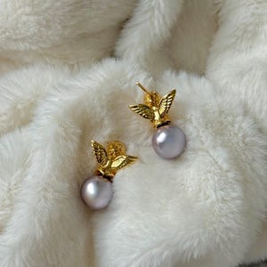 May include: A pair of gold-colored angel earrings with pearl-like, light purple orbs. The earrings are set against a white, fluffy background.