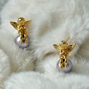 May include: A pair of gold-colored angel earrings. Each earring features a small, detailed angel figure with outstretched wings, perched atop a gray pearl. The earrings are set against a soft, white, fluffy background.