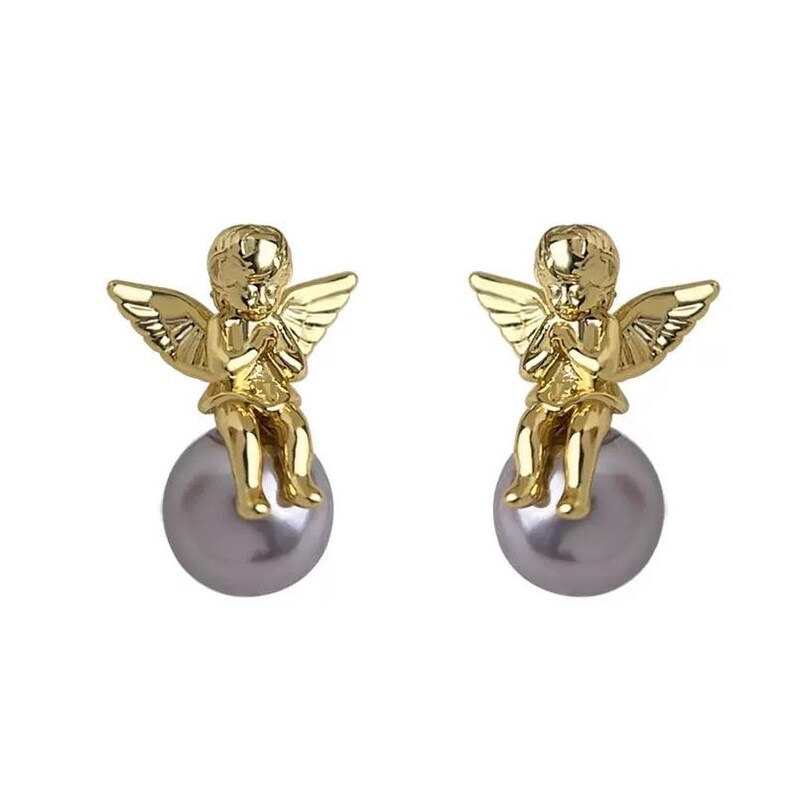 May include: A pair of gold-colored angel earrings. Each earring features a small angel figure with outstretched wings atop a gray pearl-like sphere. The earrings have a polished finish.
