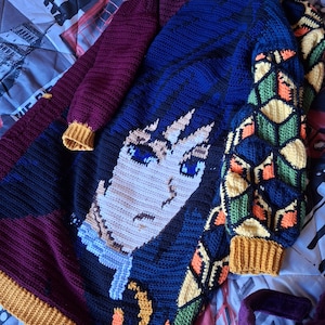 Puede incluir: Un suéter de ganchillo hecho a mano con un diseño de personaje con paneles azules, negros y burdeos. Las mangas tienen patrones geométricos en verde, naranja y amarillo. El suéter tiene un dobladillo y puños acanalados amarillos.