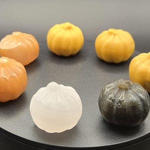 Può includere: Una collezione di sette sculture in pietra scolpite che assomigliano a piccole zucche o zucche. Le zucche sono nei toni dell'arancione, giallo, bianco e grigio scuro, disposte su una superficie circolare nera.