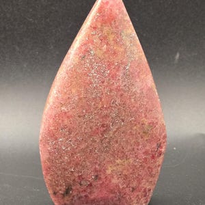 Pu&ograve; includere: Una pietra lucida a forma di goccia con una parte superiore appuntita. La pietra &egrave; principalmente di colore rosa intenso, con aree di rosa pi&ugrave; chiaro e alcune inclusioni marroni e nere. La superficie ha una consistenza leggermente ruvida.