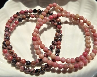 May include: Three strands of pink and brown beaded bracelets. The beads range in colour from light pink to dark brown, with some beads having a speckled appearance. The bracelets are arranged in a circular pattern on a white, scalloped dish.