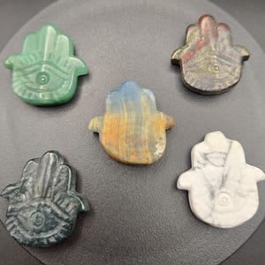 Può includere: Cinque ciondoli a forma di mano di Hamsa in pietra scolpita di vari colori, tra cui verde, blu, marrone e bianco. Ogni ciondolo presenta un disegno a occhio al centro del palmo. I ciondoli sono esposti su una superficie grigio scuro.