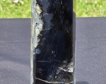 Könnte beinhalten: Ein hoher, rechteckiger schwarzer Kristallobelisk mit weißer Kappe. Der polierte Stein hat eine glänzende Oberfläche mit weißen und gelben Adern. Der Obelisk wird auf einer dunkelgrauen Oberfläche mit grünem Gras im Hintergrund präsentiert.