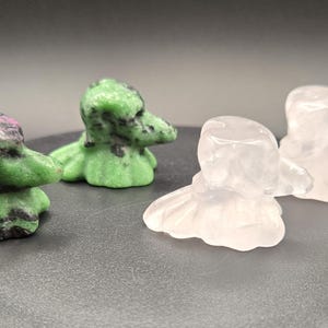Peut inclure: Quatre figurines en forme de canard sculptées dans des pierres précieuses. Deux sont vertes avec des mouchetures noires, une est un mélange de vert, de noir et de rose, et les deux autres sont transparentes. Sur une surface gris foncé.