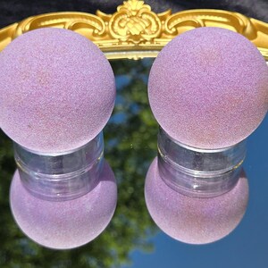 May include: Two lavender-colored bath bombs sit atop clear jars, reflected in a mirror. The bath bombs have a textured surface and are spherical. The mirror is framed by a gold-colored ornate border.