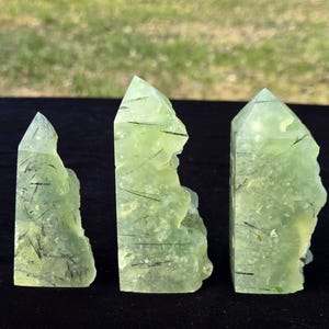 Può includere: Tre torri di cristallo di prehnite verde chiaro con inclusioni di tormalina nera. I cristalli hanno una consistenza ruvida e naturale e sono esposti su una superficie scura. I cristalli variano per dimensioni e forma, con punte appuntite.